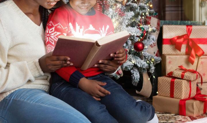 woman and child reading book in front of christmas tree and presents.